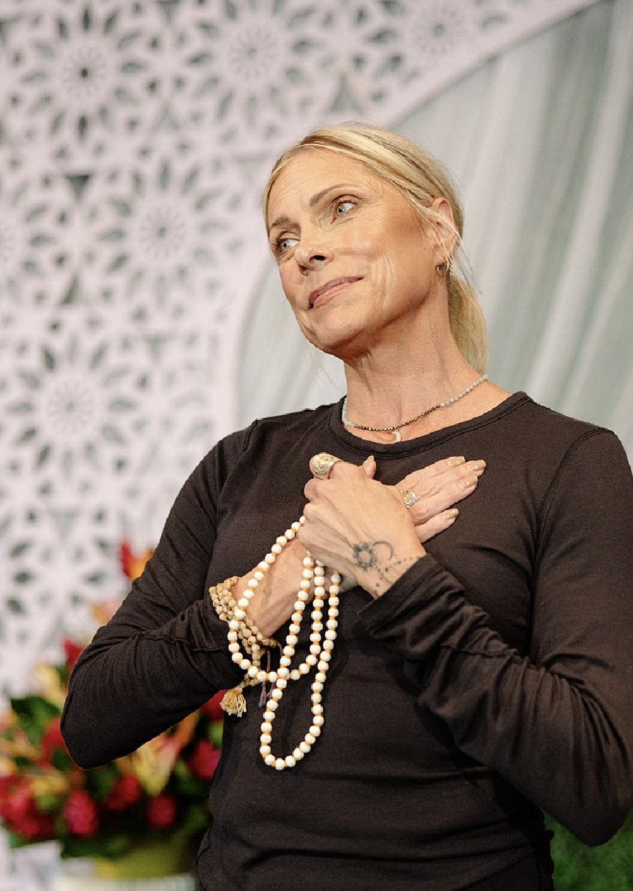 Eva Maurice on stage with a hand over her heart, holding mala beads, in front of a white lattice backdrop